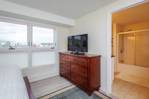 Bedroom with large window, chest of drawers with TV, and open door to a bathroom with a shower.