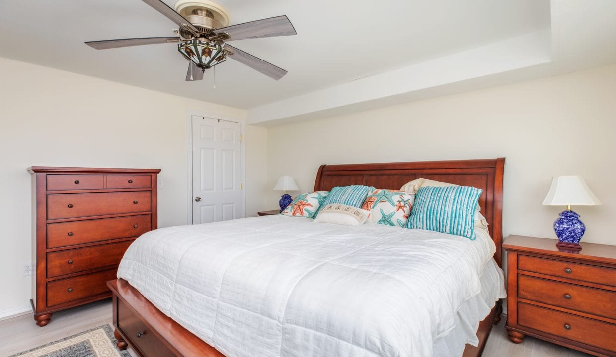 Bedroom with a large bed, colorful pillows, wooden dresser, and two matching nightstands with blue lamps. A ceiling fan is above, and a door is closed in the background.