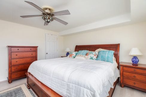 Bedroom with a large bed, colorful pillows, wooden dresser, and two matching nightstands with blue lamps. A ceiling fan is above, and a door is closed in the background.