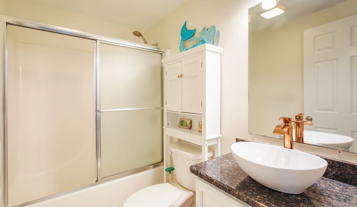 Bathroom with shower, toilet, and sink. Features a granite countertop, white cabinets, round vessel sink, and brushed bronze faucet. Overhead mirror and small decorative items present.