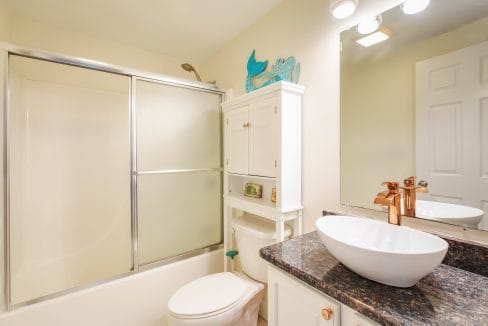 Bathroom with shower, toilet, and sink. Features a granite countertop, white cabinets, round vessel sink, and brushed bronze faucet. Overhead mirror and small decorative items present.