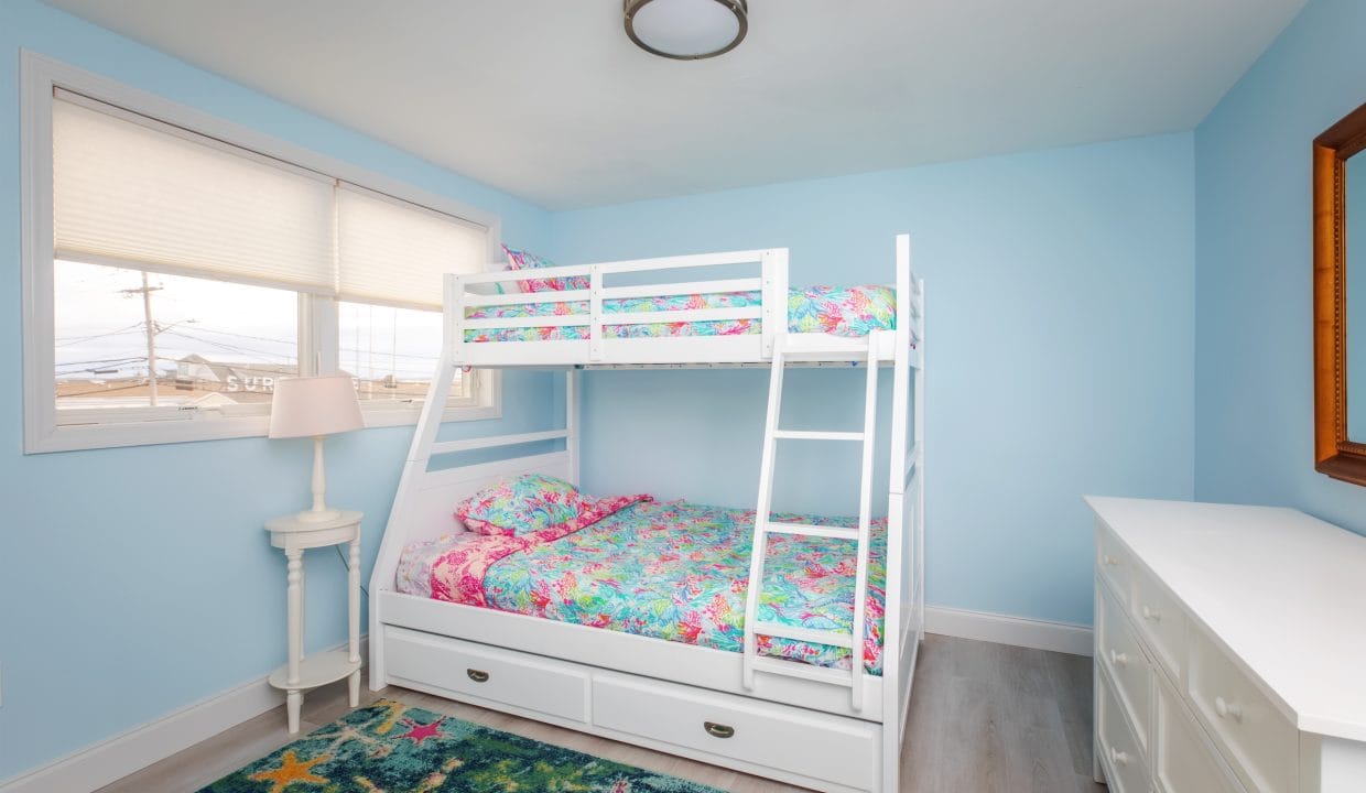 A small bedroom with a blue wall features a white bunk bed with colorful floral bedding, a white dresser, a starfish-patterned rug, and a window with a view of rooftops.