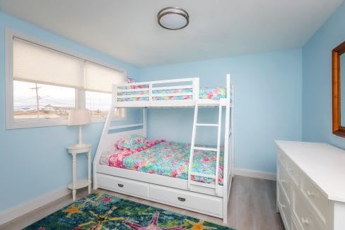 A small bedroom with a blue wall features a white bunk bed with colorful floral bedding, a white dresser, a starfish-patterned rug, and a window with a view of rooftops.