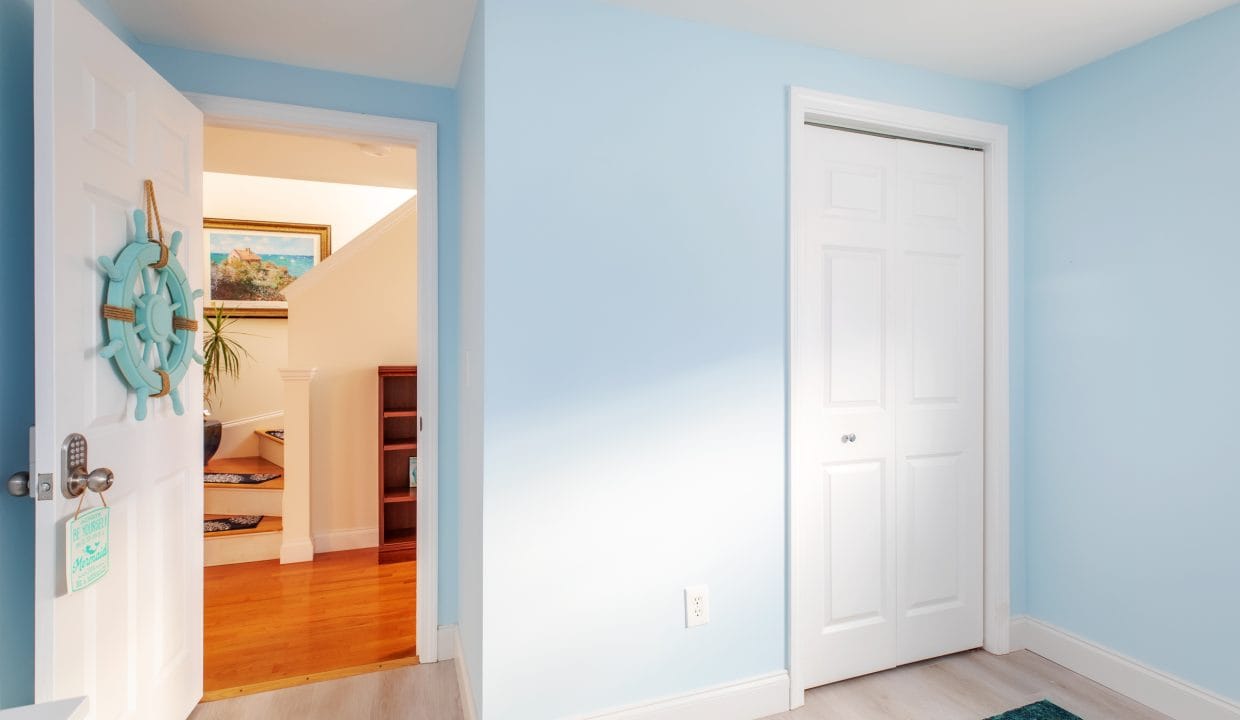 A bright room with light blue walls features a closed closet and an open door leading to a hallway with wooden flooring. A ship wheel decorates the open door.