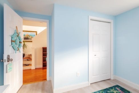 A bright room with light blue walls features a closed closet and an open door leading to a hallway with wooden flooring. A ship wheel decorates the open door.