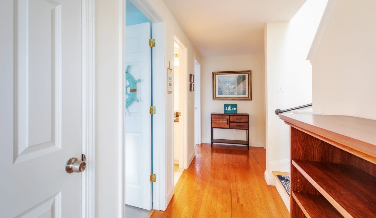 A hallway with wooden flooring, a white door on the left, a small wooden console table, and a framed picture on the wall.