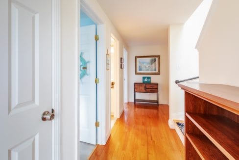 A hallway with wooden flooring, a white door on the left, a small wooden console table, and a framed picture on the wall.