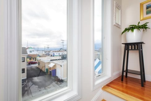 View through a large window showing a neighborhood with houses and utility poles under a cloudy sky. A tall plant on a wooden stand is positioned by the window.