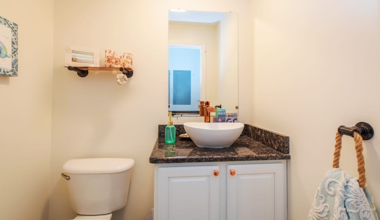 Small bathroom with a white toilet, a granite countertop with a white vessel sink, a rectangular mirror, and a wall shelf with decorative items above the toilet.