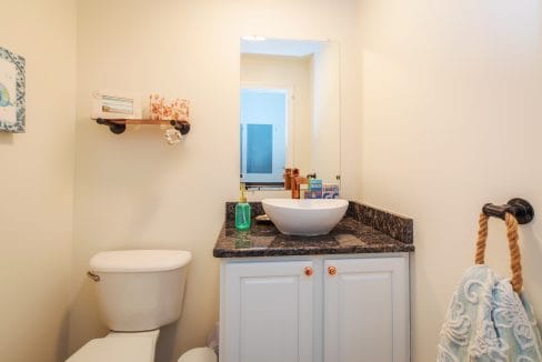 Small bathroom with a white toilet, a granite countertop with a white vessel sink, a rectangular mirror, and a wall shelf with decorative items above the toilet.