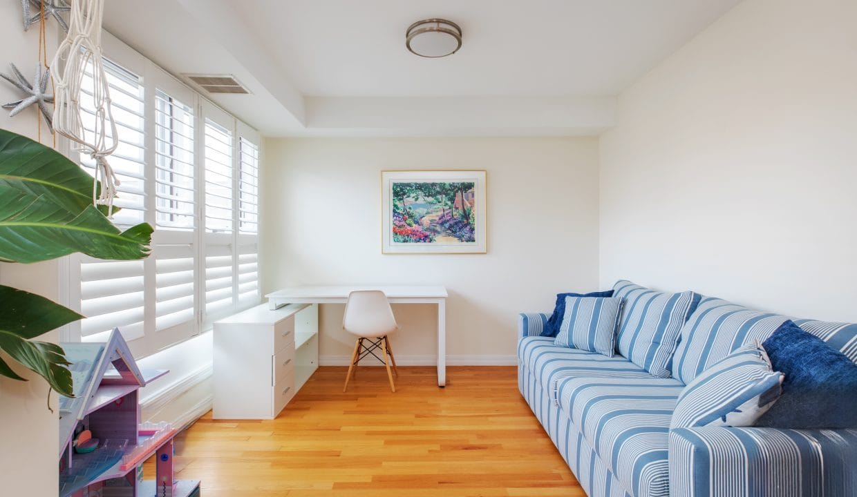 A bright, minimalist room with a striped blue sofa, a small desk and chair, a plant, a dollhouse, and a framed painting on the wall. Wood flooring and large windows with shutters.