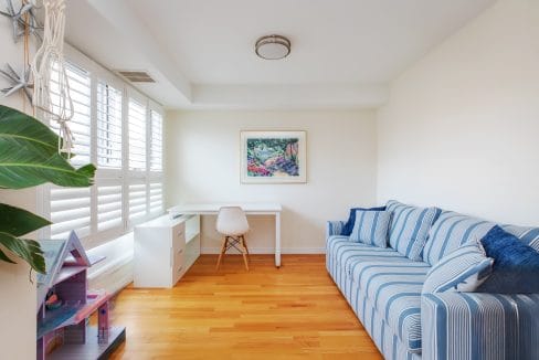 A bright, minimalist room with a striped blue sofa, a small desk and chair, a plant, a dollhouse, and a framed painting on the wall. Wood flooring and large windows with shutters.