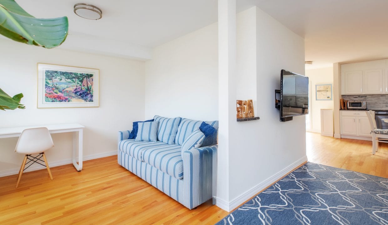 A living room with a striped blue sofa, a wall-mounted TV, a small white desk with a chair, and a blue-patterned rug on wooden flooring. A kitchen area is visible in the background.