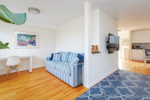 A living room with a striped blue sofa, a wall-mounted TV, a small white desk with a chair, and a blue-patterned rug on wooden flooring. A kitchen area is visible in the background.