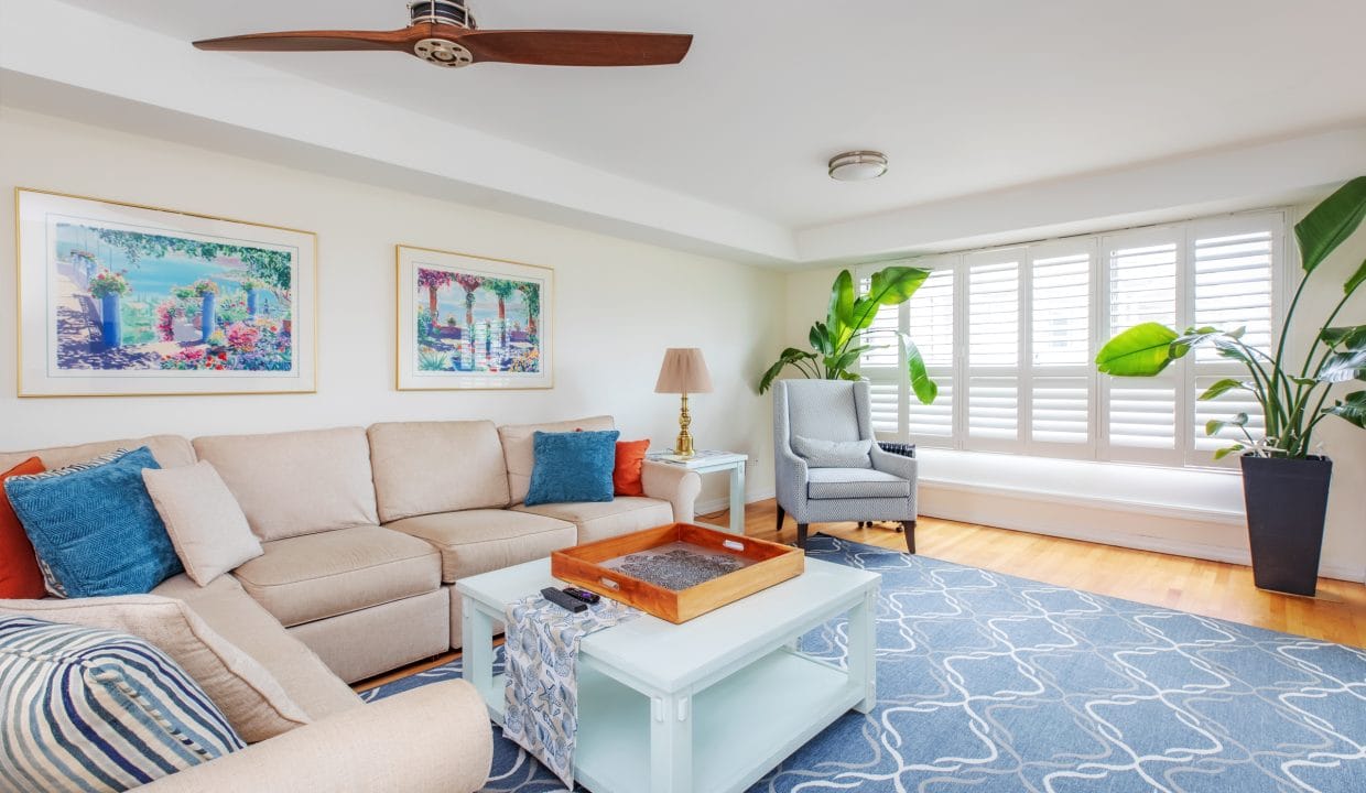 A bright living room with a beige sectional sofa, blue and white patterned rug, two framed artworks, a ceiling fan, and large plants near a window with white shutters.