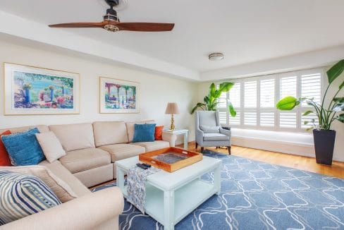 A bright living room with a beige sectional sofa, blue and white patterned rug, two framed artworks, a ceiling fan, and large plants near a window with white shutters.