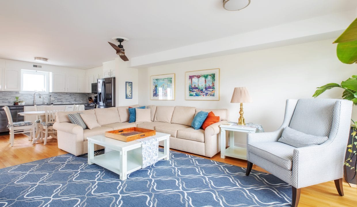 Spacious living room with a beige sectional sofa, blue patterned rug, and white coffee table. There's a gray armchair, a lamp, and wall art. A kitchen and dining area are visible in the background.