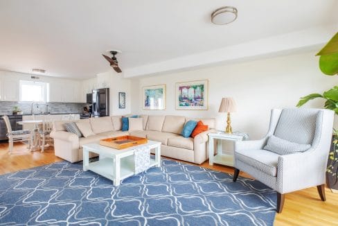 Spacious living room with a beige sectional sofa, blue patterned rug, and white coffee table. There's a gray armchair, a lamp, and wall art. A kitchen and dining area are visible in the background.