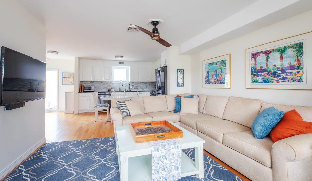 Modern living room with beige sectional sofa, blue and orange cushions, wall art, and a ceiling fan. A large TV is mounted on the wall. The kitchen is visible in the background.