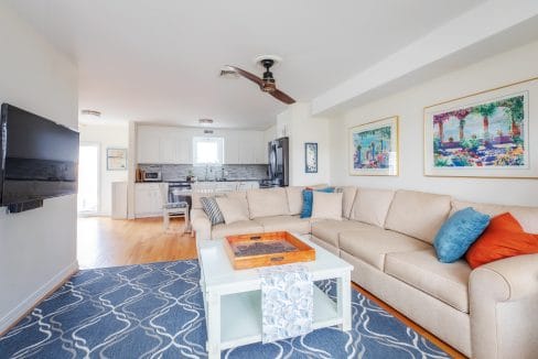 Modern living room with beige sectional sofa, blue and orange cushions, wall art, and a ceiling fan. A large TV is mounted on the wall. The kitchen is visible in the background.