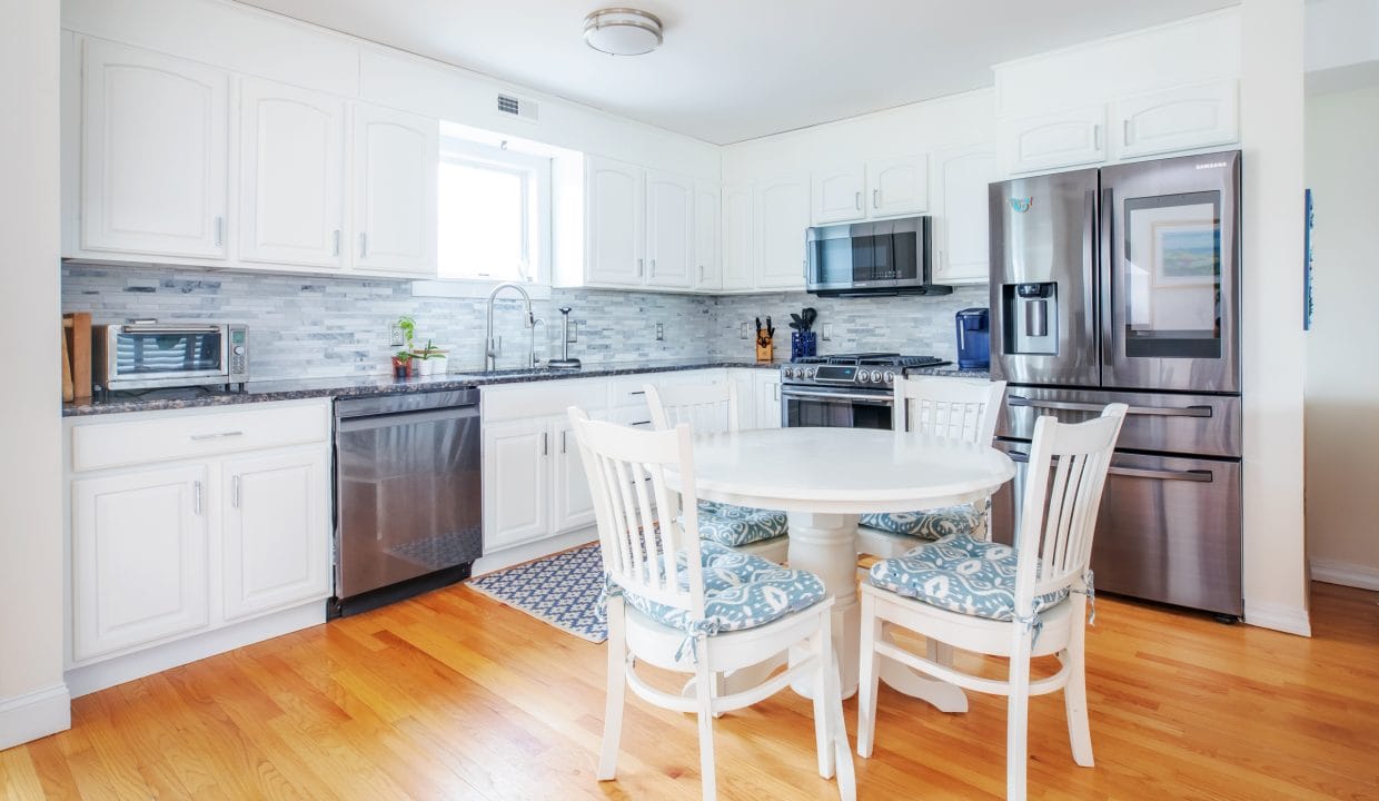 Modern kitchen with white cabinets, stainless steel appliances, round dining table, and hardwood floor. A window provides natural light above the sink.