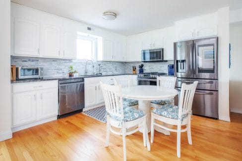 Modern kitchen with white cabinets, stainless steel appliances, round dining table, and hardwood floor. A window provides natural light above the sink.