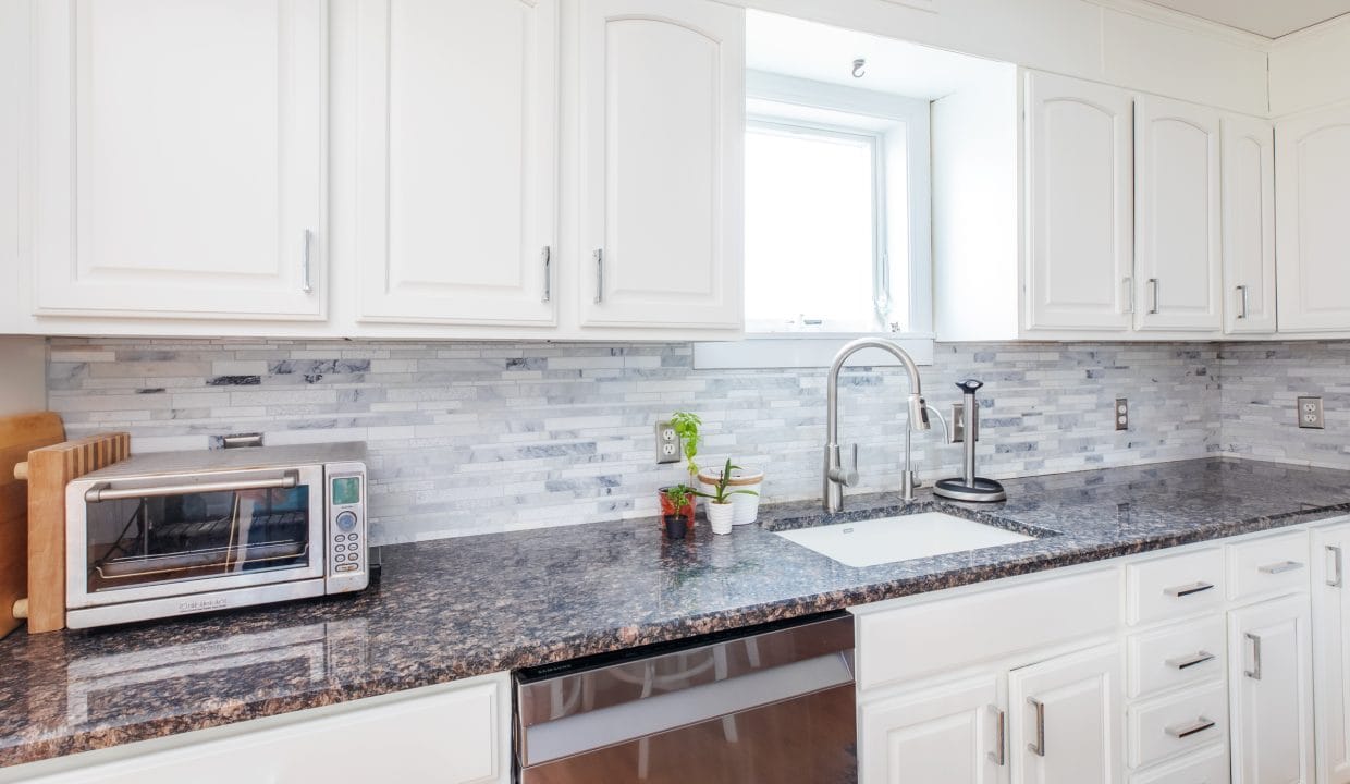 A modern kitchen with white cabinets, a dark granite countertop, a toaster oven, a sink, and a window above. Light gray tile backsplash adds contrast.