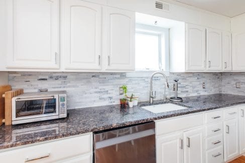 A modern kitchen with white cabinets, a dark granite countertop, a toaster oven, a sink, and a window above. Light gray tile backsplash adds contrast.