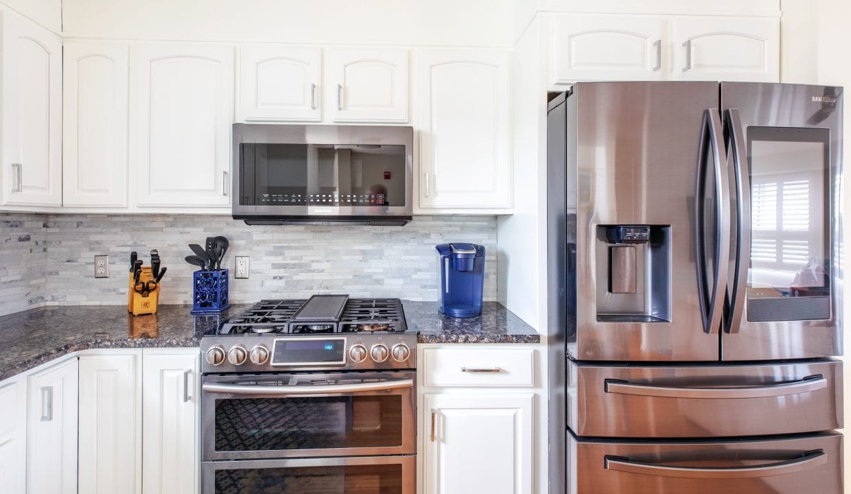 Modern kitchen with white cabinets, stainless steel refrigerator, double oven, microwave, knife block, and a blue water filter on the counter.