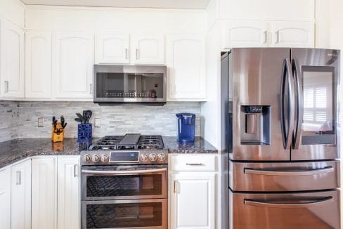 Modern kitchen with white cabinets, stainless steel refrigerator, double oven, microwave, knife block, and a blue water filter on the counter.