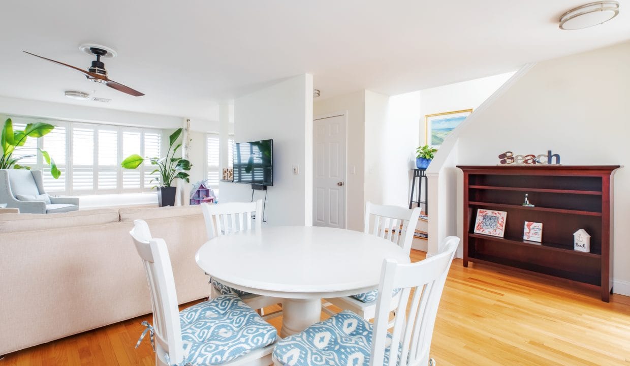 Bright, airy living room with a round dining table and four chairs, a ceiling fan, light wood flooring, large windows with shutters, a TV, and a bookshelf with decor.