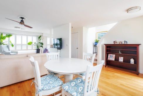 Bright, airy living room with a round dining table and four chairs, a ceiling fan, light wood flooring, large windows with shutters, a TV, and a bookshelf with decor.