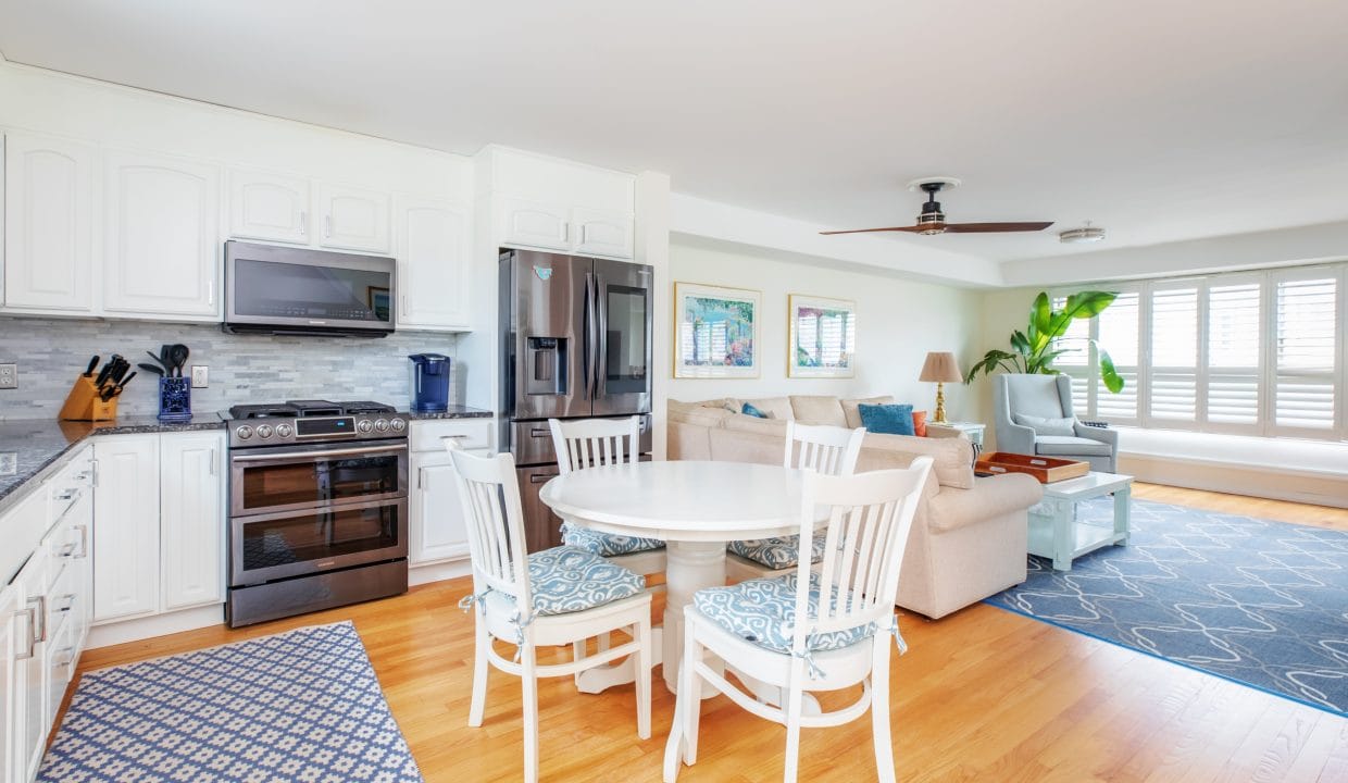 Open-concept living space with a kitchen featuring white cabinets, a round dining table with four chairs, and a living area with a sofa, ceiling fan, and large window with shutters.