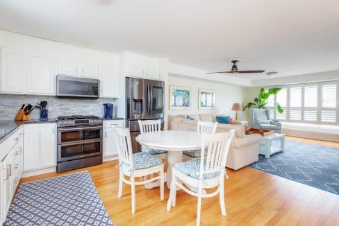 Open-concept living space with a kitchen featuring white cabinets, a round dining table with four chairs, and a living area with a sofa, ceiling fan, and large window with shutters.