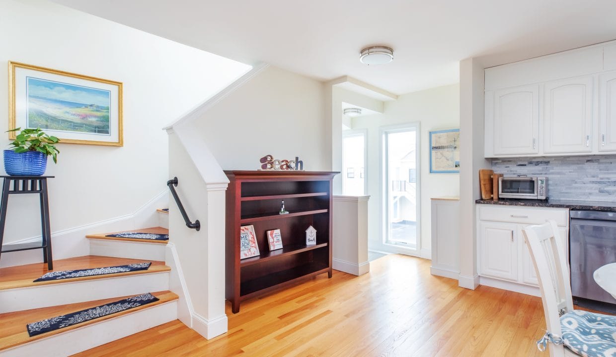 A bright kitchen with wooden floors, white cabinets, and a small dining table. A staircase with carpeting leads upstairs. A bookshelf and a plant are near the entrance.