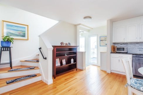 A bright kitchen with wooden floors, white cabinets, and a small dining table. A staircase with carpeting leads upstairs. A bookshelf and a plant are near the entrance.