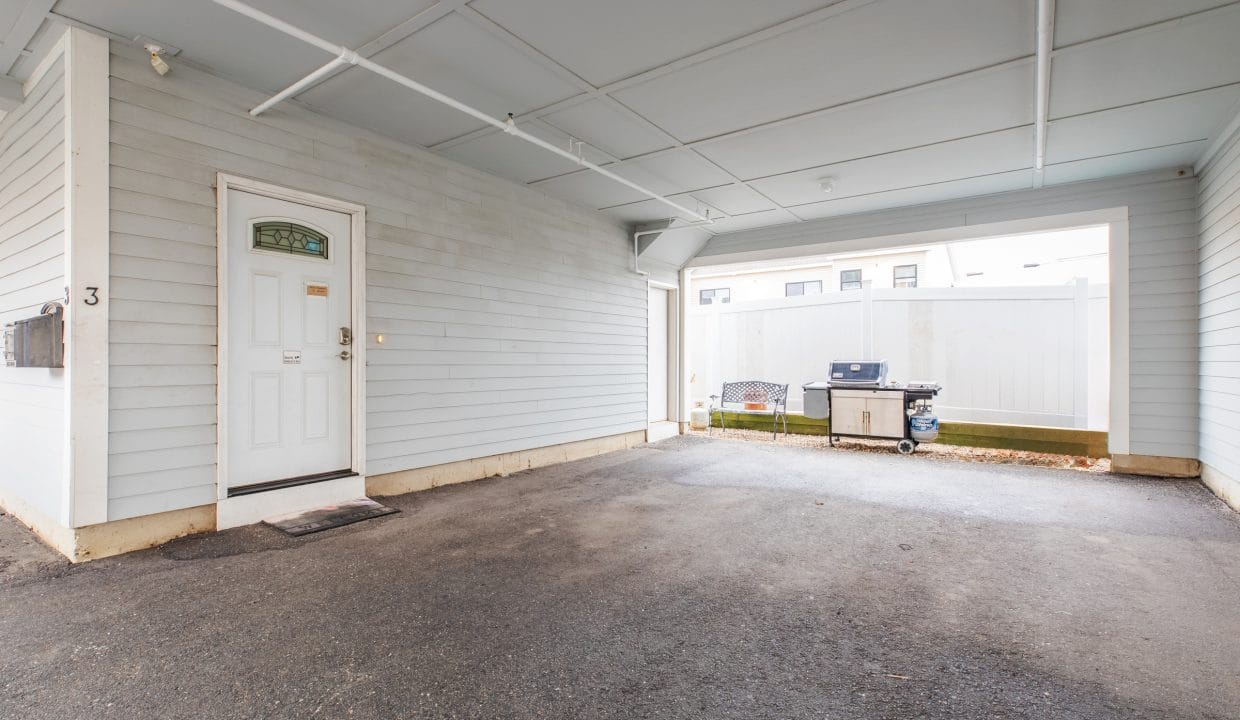 Covered parking area with a closed white door on the left, a barbecue grill, and a bench on the right. The area has a light blue exterior and ceiling with white support beams.