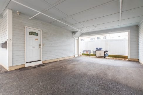 Covered parking area with a closed white door on the left, a barbecue grill, and a bench on the right. The area has a light blue exterior and ceiling with white support beams.