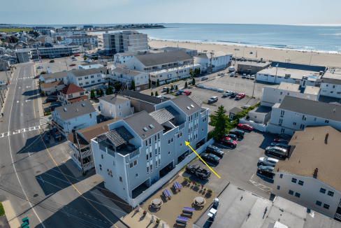 Aerial view of a coastal town with several buildings, a beach, and parking lots. A yellow arrow points to a specific building.