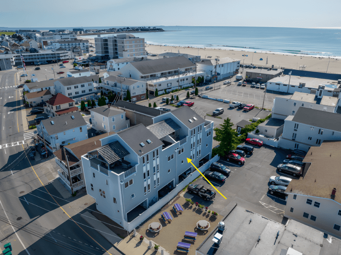 Aerial view of a coastal town with several buildings, a beach, and parking lots. A yellow arrow points to a specific building.