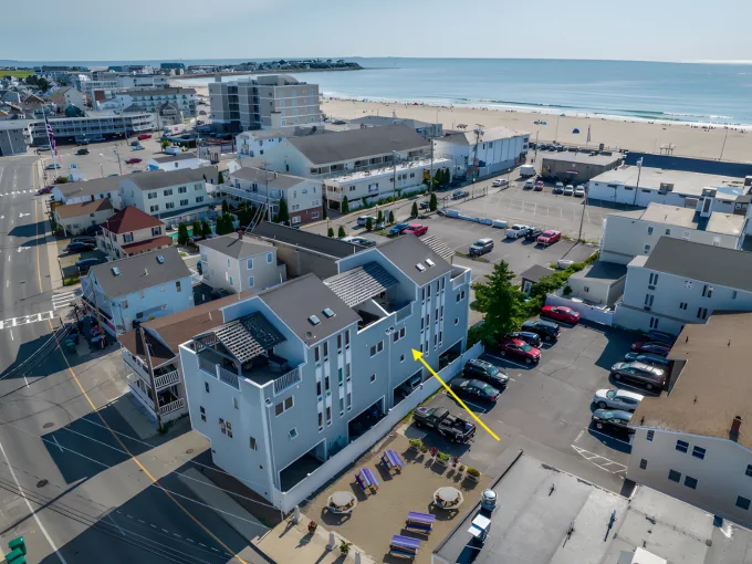 Aerial view of a coastal town with several buildings, a beach, and parking lots. A yellow arrow points to a specific building.