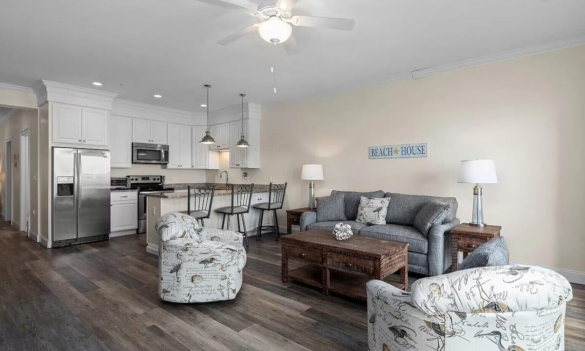 Open-plan living room and kitchen with wooden floors, gray sofa, two patterned armchairs, ceiling fan, and stainless steel kitchen appliances. Wall sign reads 