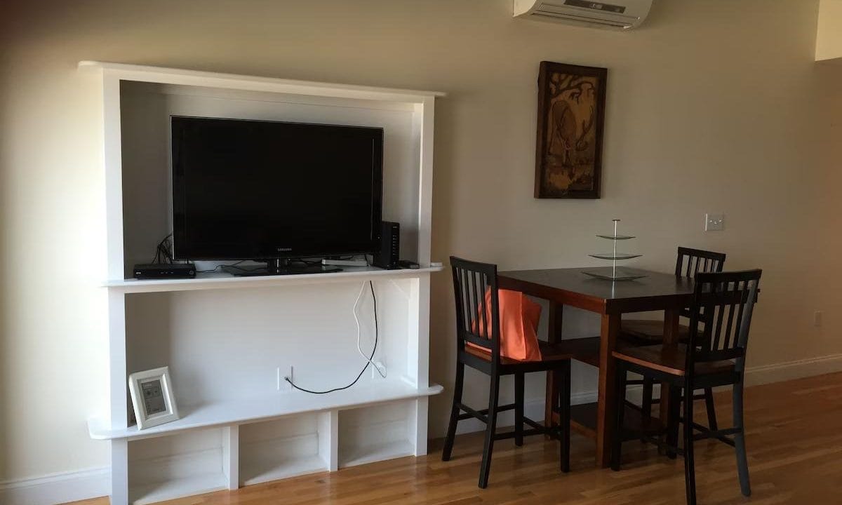 Living room with a wall-mounted TV stand, a TV, and a small dining table with four chairs. Air conditioner on wall above table, and picture frame below TV.