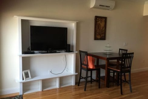 Living room with a wall-mounted TV stand, a TV, and a small dining table with four chairs. Air conditioner on wall above table, and picture frame below TV.