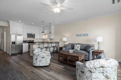 Open-plan living room and kitchen with wooden floors, gray sofa, two patterned armchairs, ceiling fan, and stainless steel kitchen appliances. Wall sign reads 