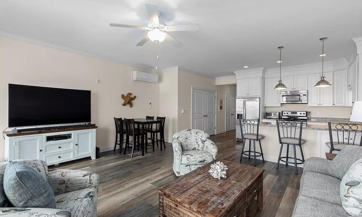 Spacious open-plan living room and kitchen with modern furnishings, including a couch, armchairs, dining table, flat-screen TV, and kitchen island with bar stools.