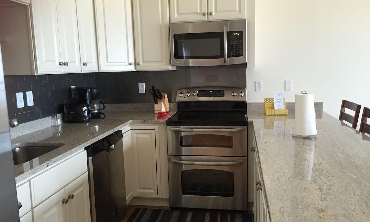 Modern kitchen with white cabinets, black backsplash, and stainless steel appliances, including a microwave and oven. Granite countertop with a roll of paper towels and a kitchen island.