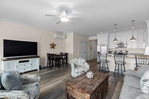 Spacious open-plan living room and kitchen with modern furnishings, including a couch, armchairs, dining table, flat-screen TV, and kitchen island with bar stools.