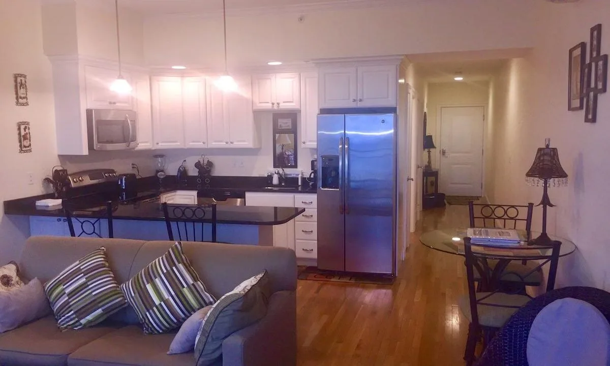 Modern kitchen and living area with stainless steel appliances, white cabinetry, and a sofa with striped pillows. Hardwood floors and a small dining table are also visible.