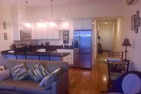 Modern kitchen and living area with stainless steel appliances, white cabinetry, and a sofa with striped pillows. Hardwood floors and a small dining table are also visible.